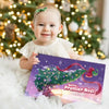 Bébé assis devant un sapin tenant le livre personnalisé “Notre Premier Noël 2025” avec Ava et Papa.
